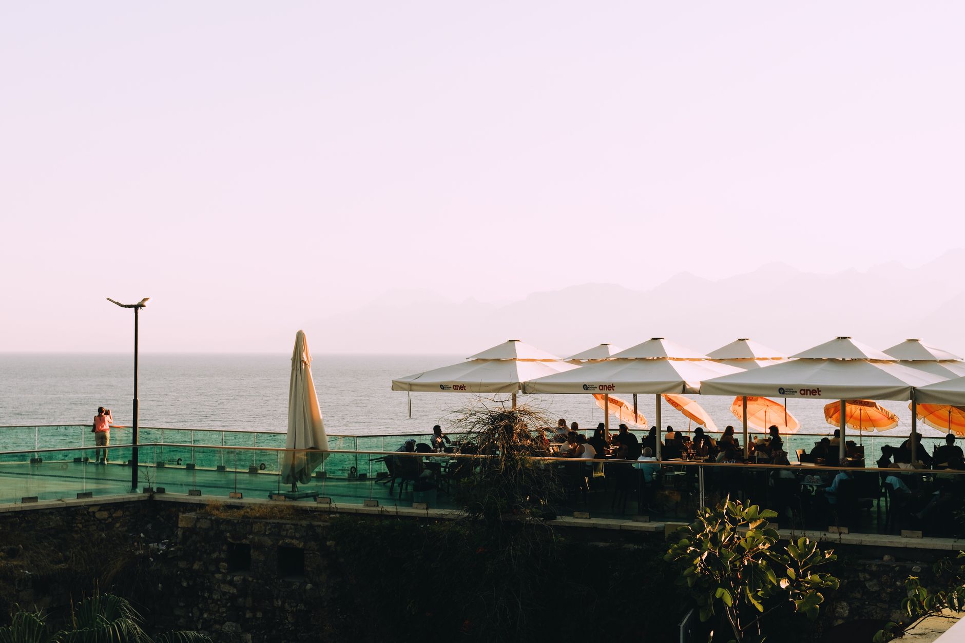 Terrasse de restaurant extérieure avec vue sur l'océan, parasols et personnes dînant au coucher du soleil.