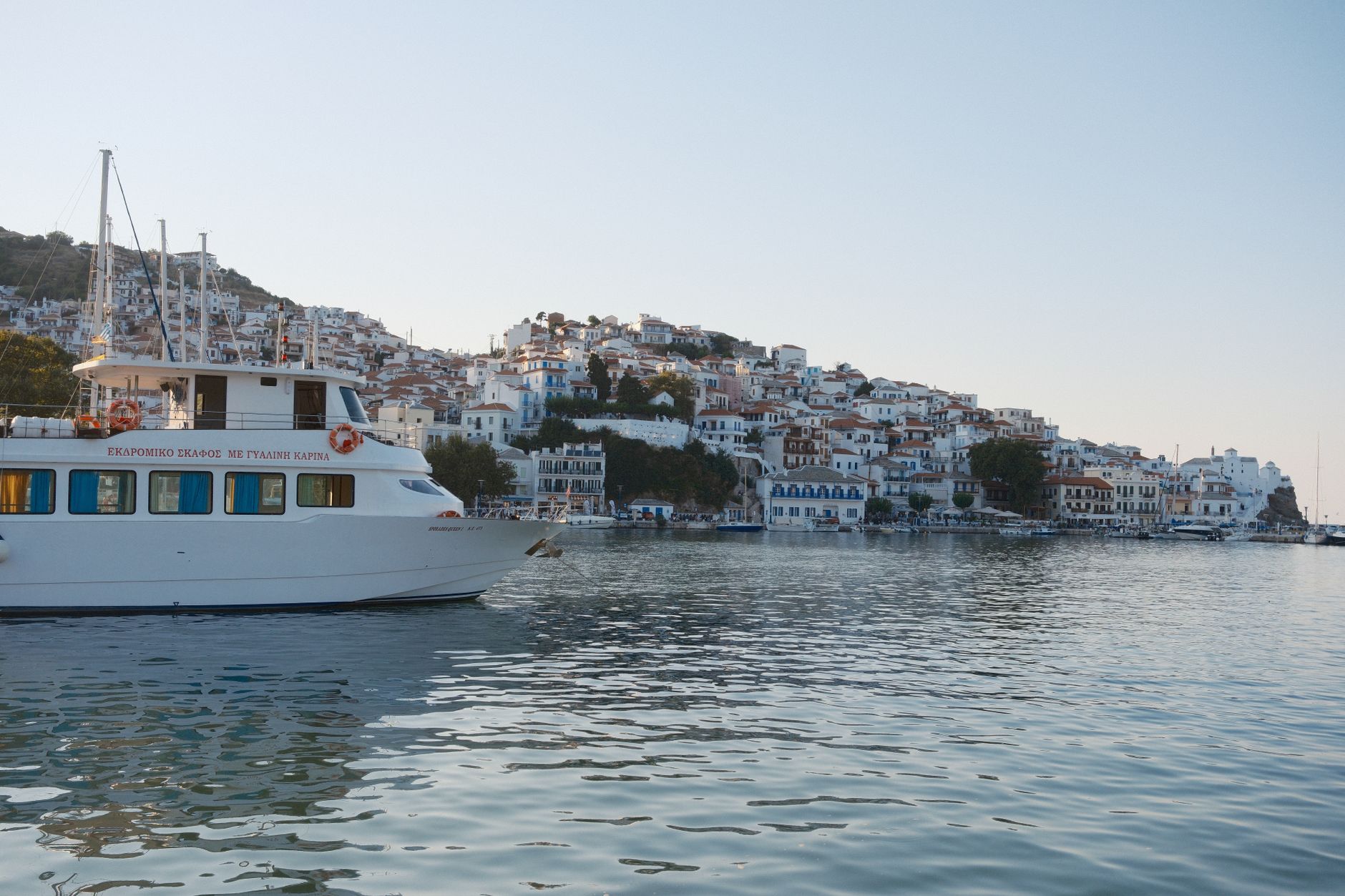 Bateau blanc sur une eau calme devant un village portuaire aux maisons blanches étagées sur une colline, sous un ciel clair.
