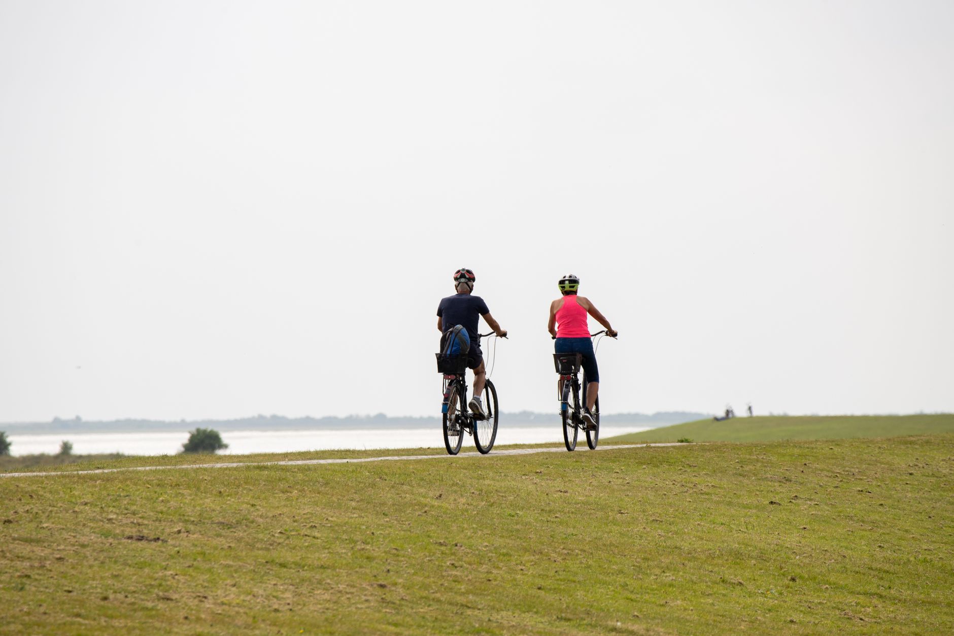 Deux cyclistes casqués, vus de dos, roulent sur un chemin au bord d’une digue herbeuse, face à une étendue d’eau sous un ciel clair.